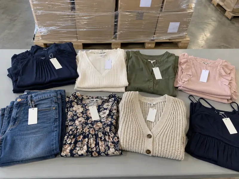 Folded women's tops, jeans, a floral dress and a knit cardigan with blank retail tags on a warehouse table, pallets of cartons behind
